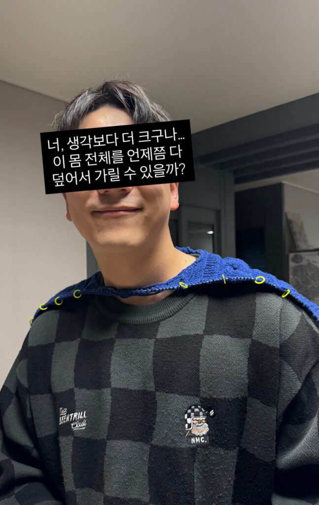 체크무늬 스웨터를 입은 남성이 작업 중인 네이비 니트 편물을 어깨에 걸쳐 사이즈를 가늠해보는 모습. 눈 부분은 검은 박스로 가려져 있고, 어깨 라인에는 노란색 스티치 마커가 꽂혀 있다.