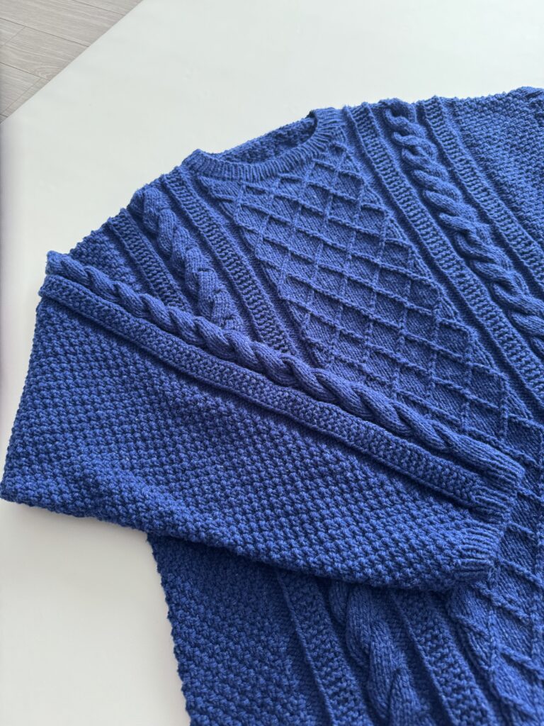 네이비 컬러 케이블 니트 스웨터가 테이블 위에 펼쳐져 있으며, 목 고무단까지 완성된 상태로 소매가 몸통 위에 가지런히 놓여 있다.