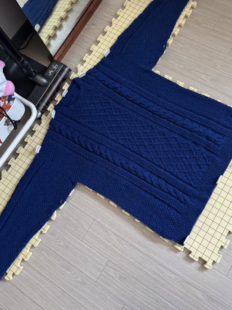 네이비 케이블 니트 스웨터를 바닥 매트 위에 펼쳐 핀으로 고정하며 블로킹 중인 모습.