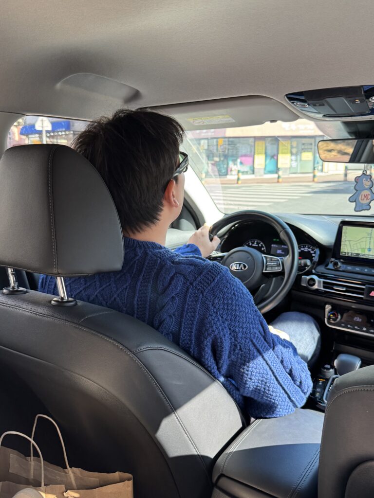 차를 운전하는 남성의 뒷모습으로, 네이비 케이블 니트 등판이 보인다.