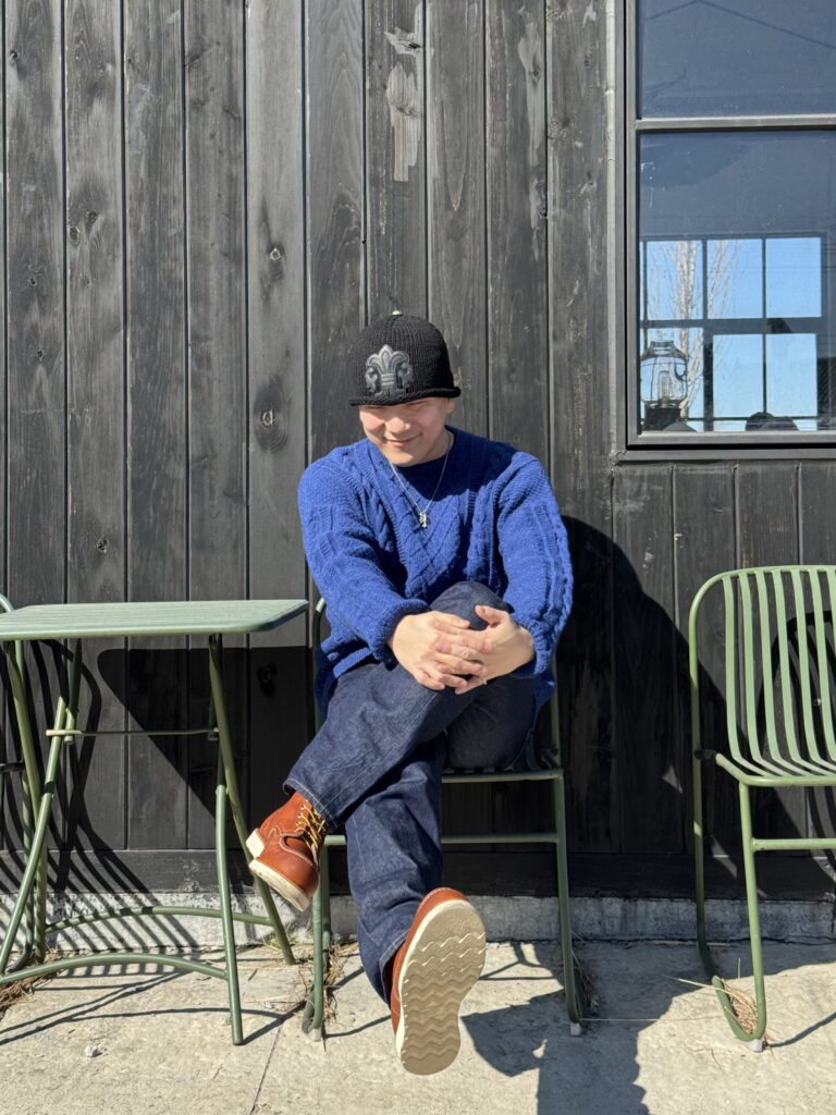 야외 의자에 앉아 다리를 꼬고 네이비 케이블 니트를 입은 남성.