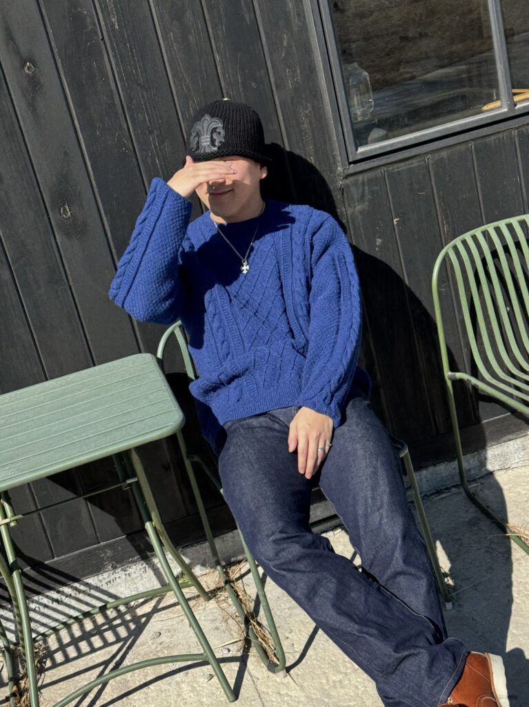 야외 카페 의자에 앉아 네이비 케이블 니트 스웨터를 입고 햇빛을 가리는 모습.