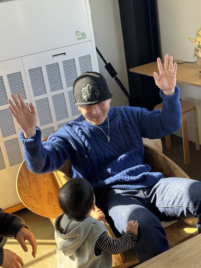 네이비 케이블 니트 스웨터를 입은 남성이 아이와 마주 보며 두 손을 들고 있는 모습.