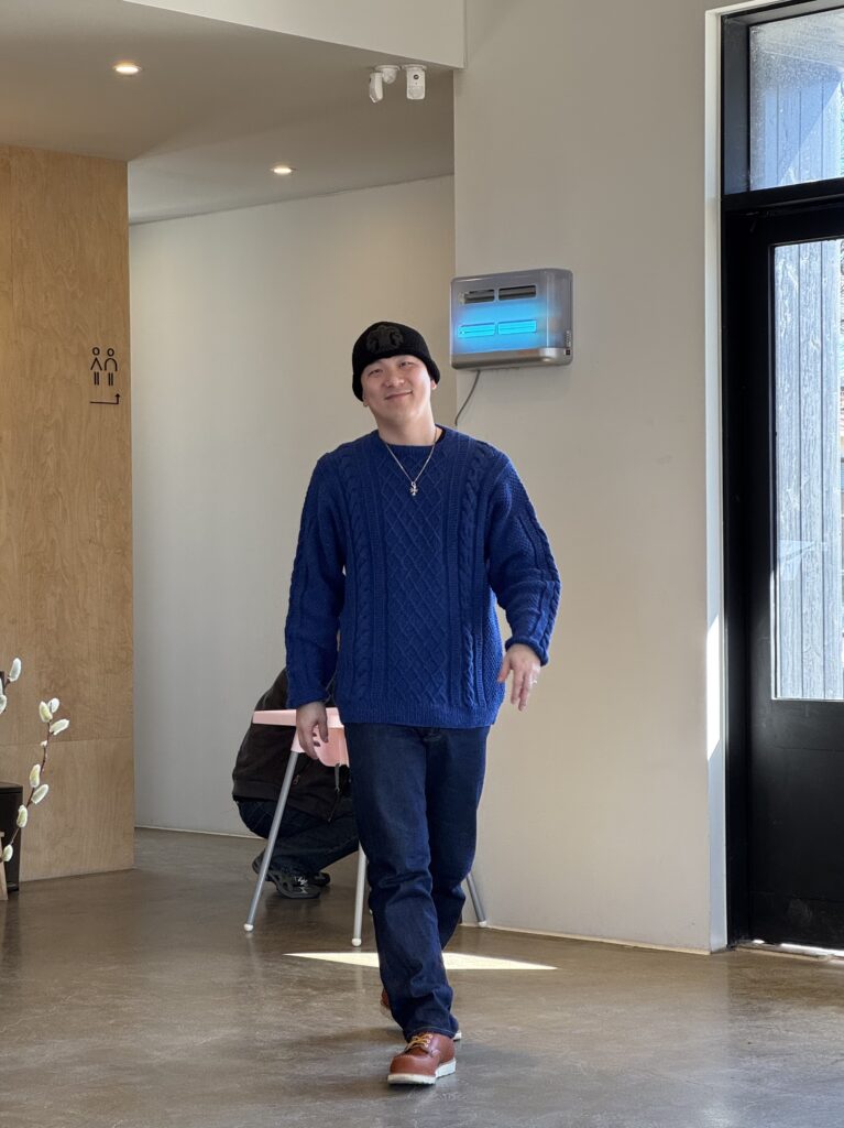 네이비 케이블 니트 스웨터를 입은 남성이 실내 공간에서 걸어오는 모습.