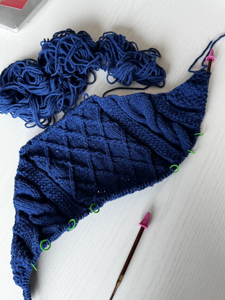 화이트 테이블 위에 놓인 네이비 컬러 케이블 니트 편물과 엉켜 있는 실, 분홍색 바늘 마개가 꽂힌 니트 바늘, 초록색 스티치 마커가 보이는 작업 중인 모습.