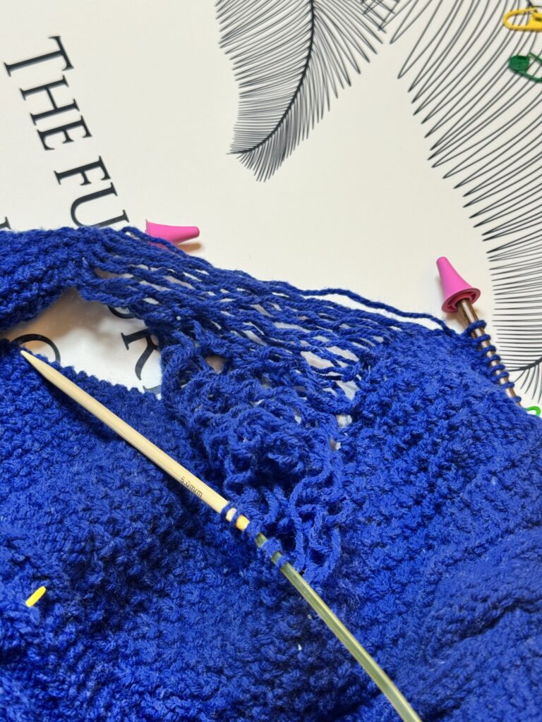 네이비 케이블 니트 스웨터의 어깨 부분을 푸르시오한 모습으로, 실이 여러 가닥으로 풀려 늘어져 있고 바늘과 분홍색 바늘 마개가 보인다.