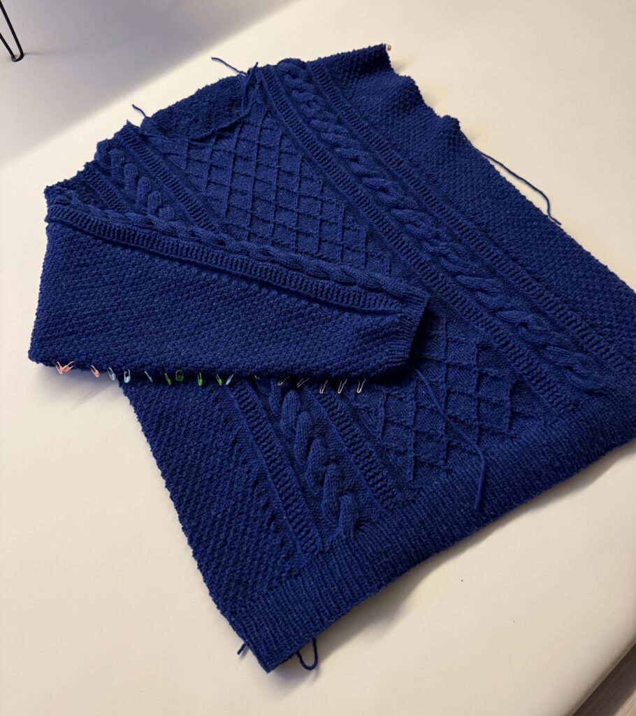 화이트 테이블 위에 펼쳐진 네이비 케이블 니트 스웨터 몸통과 완성된 소매 한 짝이 겹쳐 놓여 있으며, 반대쪽 소매를 뜨기 위해 코를 주울 부분에 스티치 마커가 꽂혀 있다.