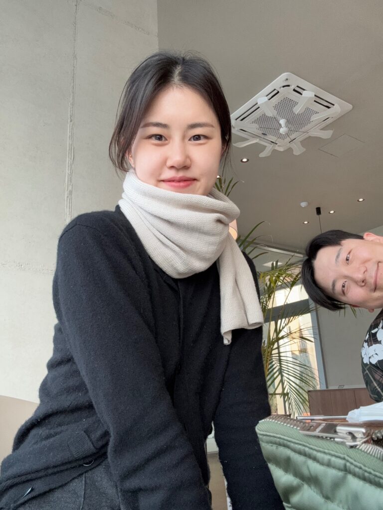 오트밀 컬러의 니트 목도리를 착용한 여성이 카페에서 미소 짓고 있는 모습